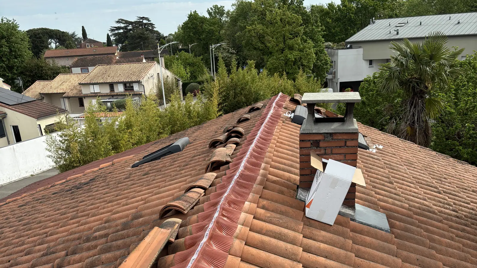 Faîtage de tuile canal en cours de pose sur closoir ventilé, souche de cheminée en brique, arbres et toits du quartier sous un ciel voilé
